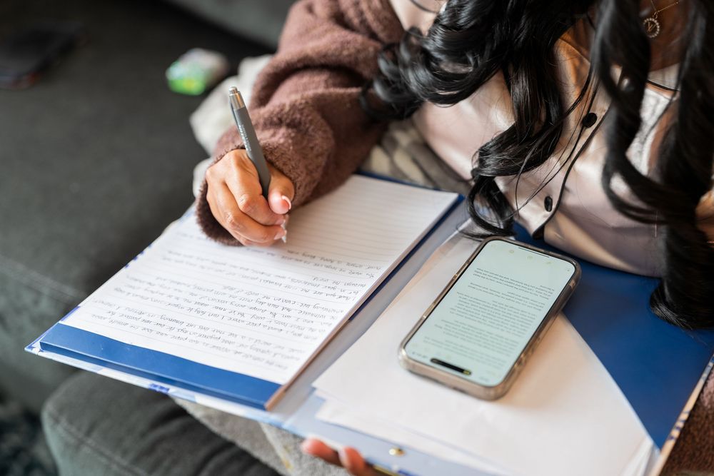 Someone writing in a notebook while studying next to a smartphone on a blue surface.