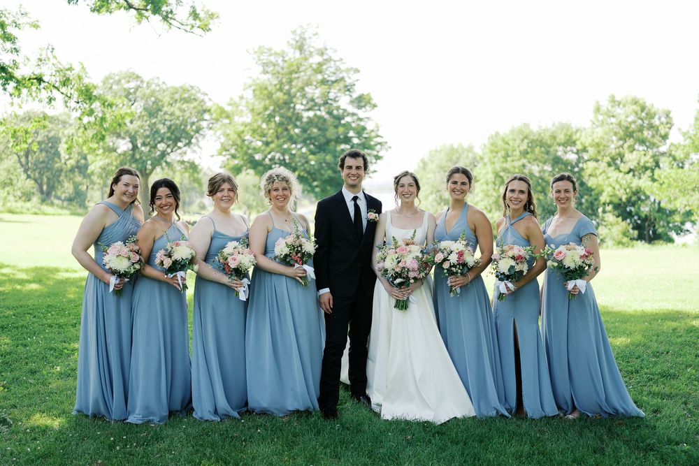 Wedding party wearing dusty blue bridesmaids dresses and black tuxedos pose together outdoors on a sunny day.
