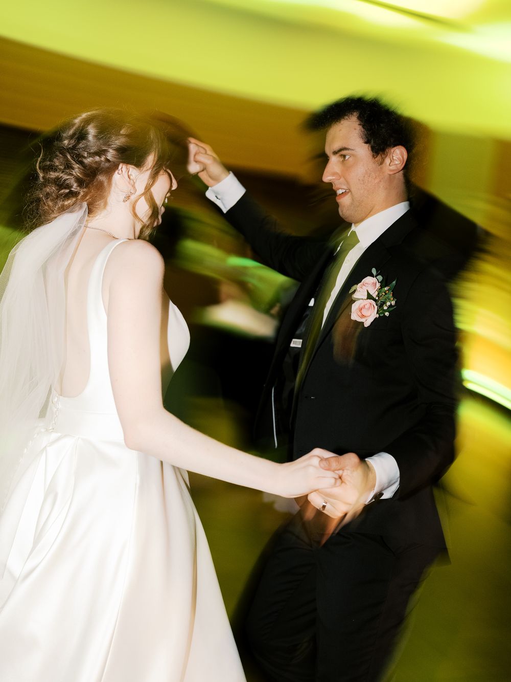 A couple in formal wedding attire dances together during their first dance at their reception under yellow lighting.