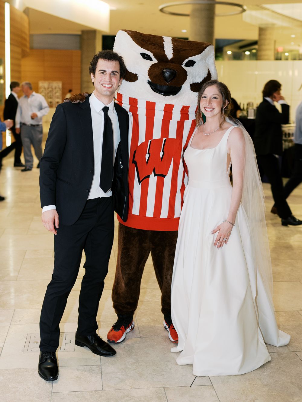 Couple in formal wedding attire walking alongside someone in a red and white striped sports mascot costume.