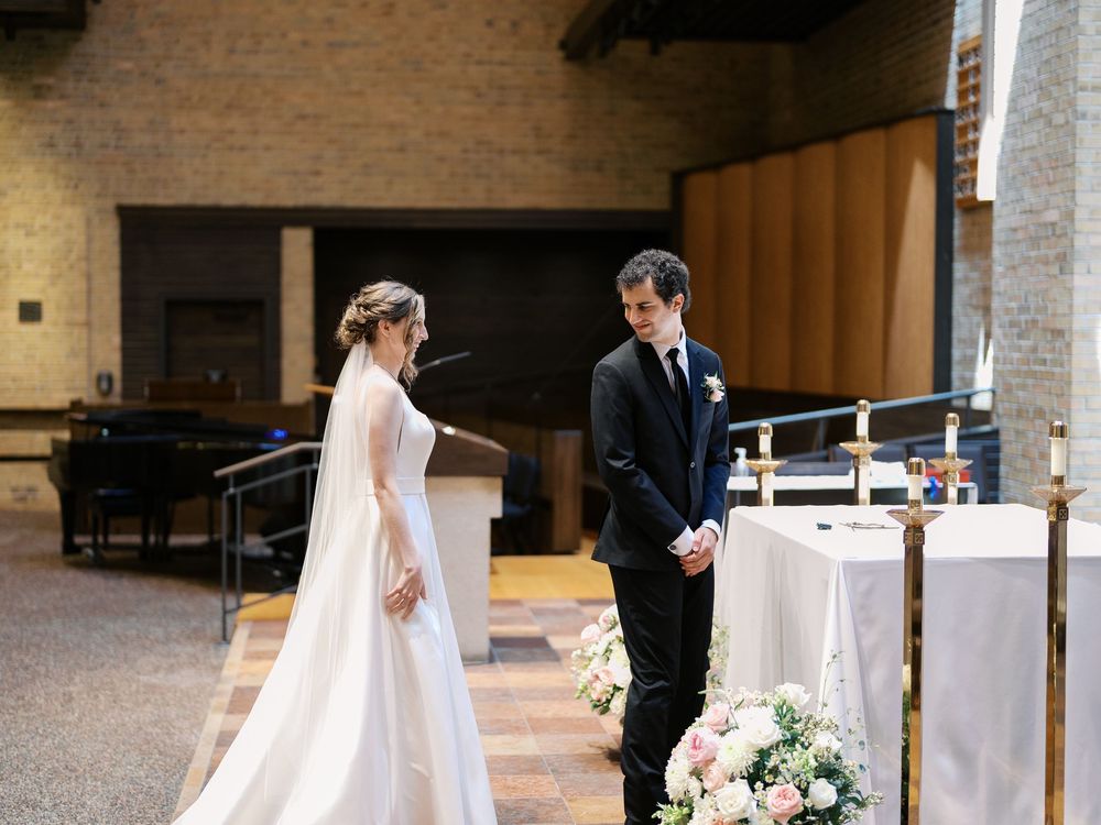 A bride and groom stand at an altar with a piano and floral arrangements in a brick-walled church venue.