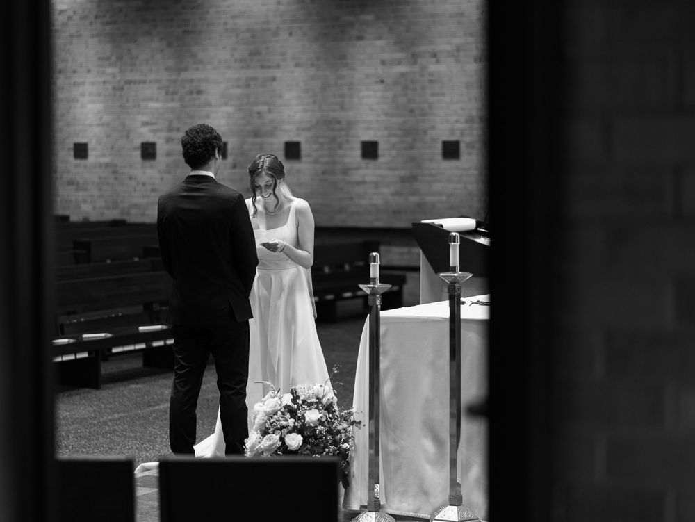 A black and white wedding ceremony photograph captured from a distance inside a church with floral arrangements.