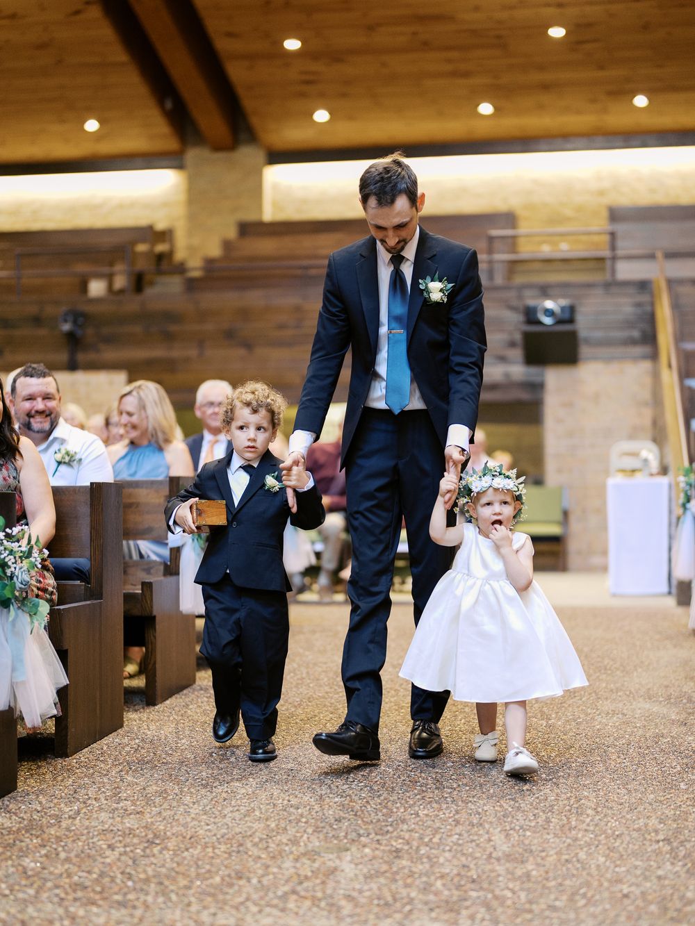 Wedding party member walks down church aisle with ring bearer in navy suit and flower girl in white dress.