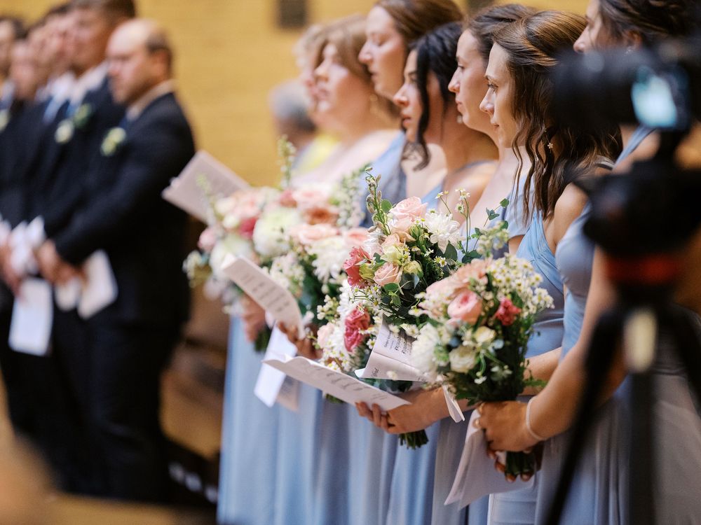 Bridesmaids in light blue dresses stand in line holding beautiful bouquets of pink and white flowers during a ceremony.