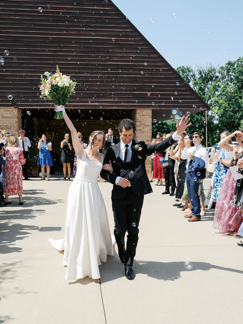 Wedding couple makes grand exit through confetti and bubbles as wedding guests celebrate outside a rustic venue.