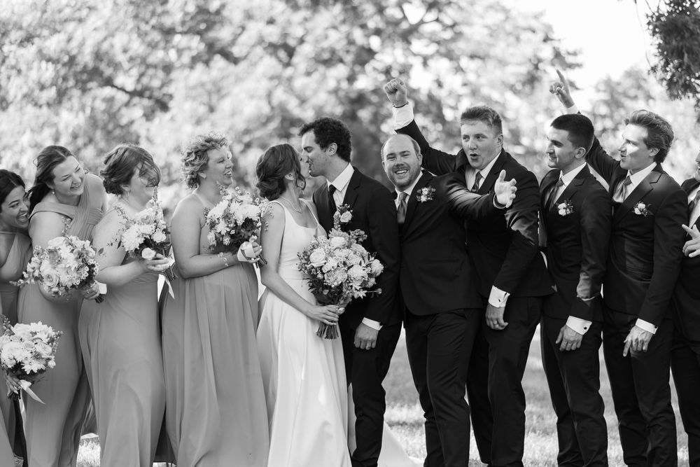 A wedding party in black and white laughs together while holding bouquets of white flowers in an outdoor setting.