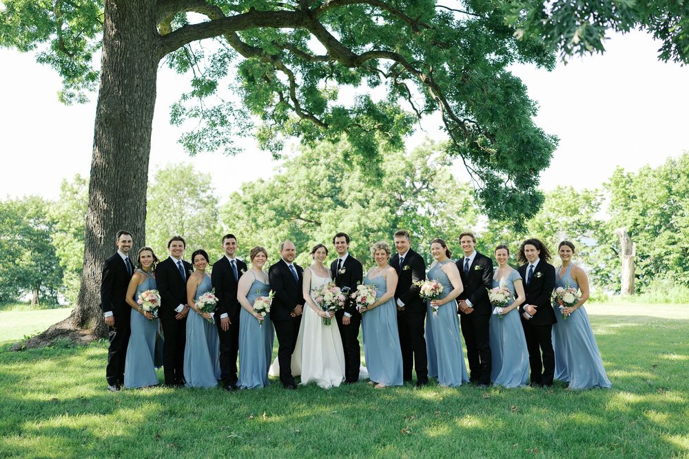 A wedding party wearing formal attire poses for photographs outdoors in a scenic park setting under green trees.