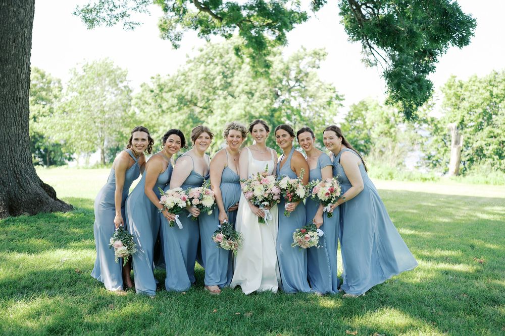 Wedding party wearing light blue bridesmaid dresses gathered outdoors under a large tree on a sunny day.