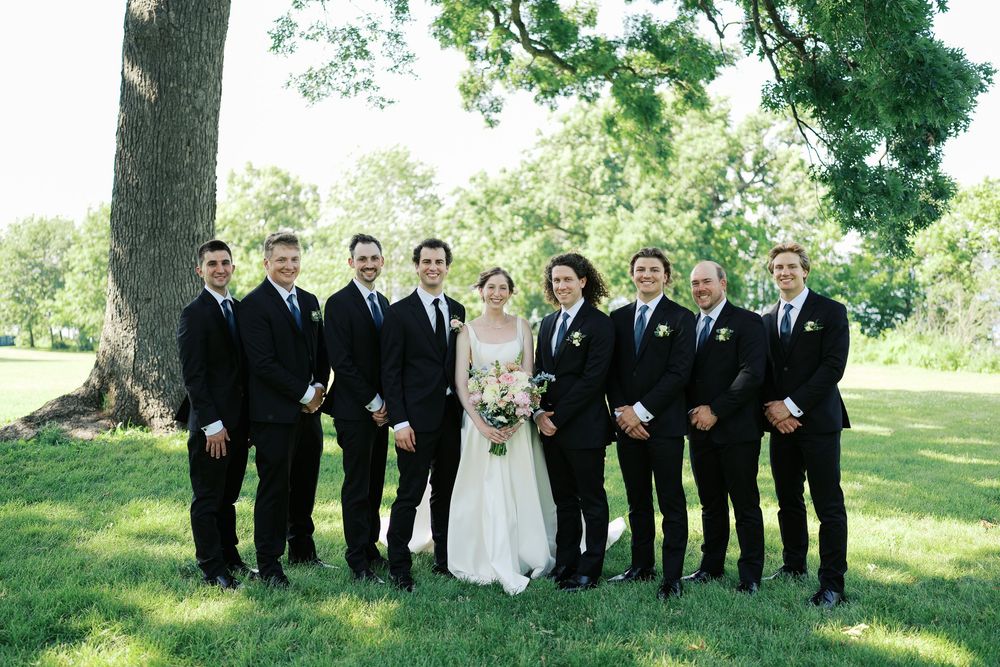 Wedding party in formal attire with black suits and white dress posing together on grass under trees.