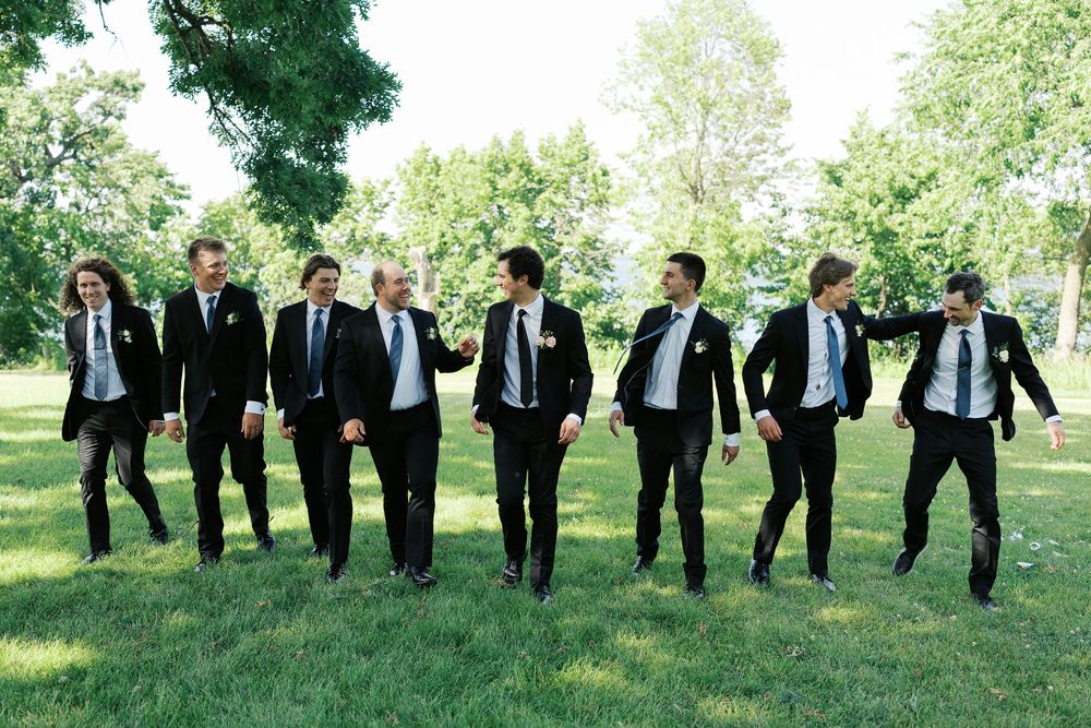 Wedding party of groomsmen in black suits and blue ties pose creatively in a line on grassy hill outdoors.