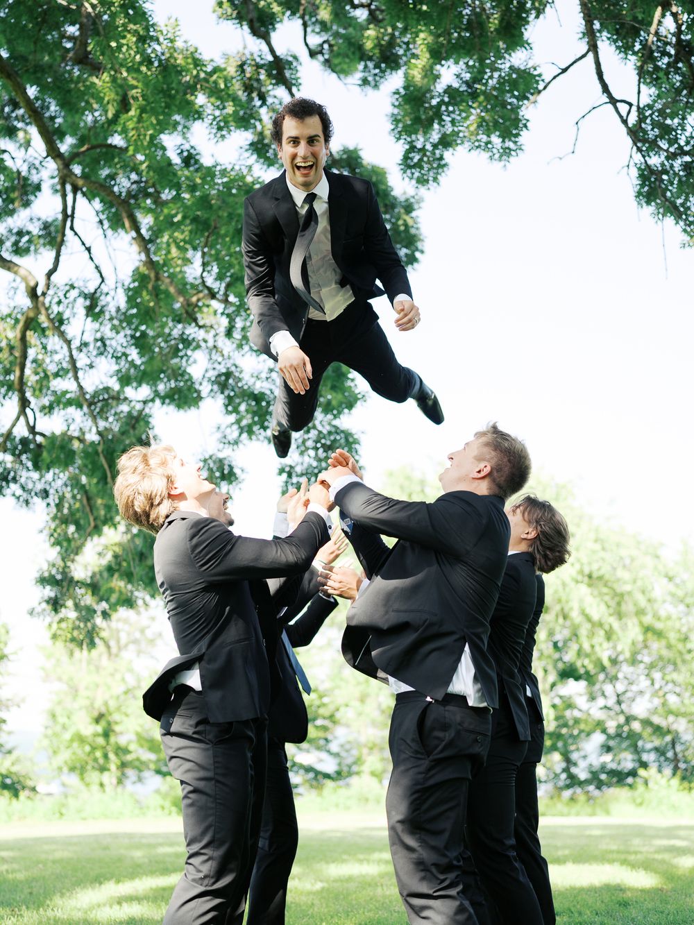 A group in formal attire performs a playful acrobatic lift in a outdoor garden setting with lush green trees.