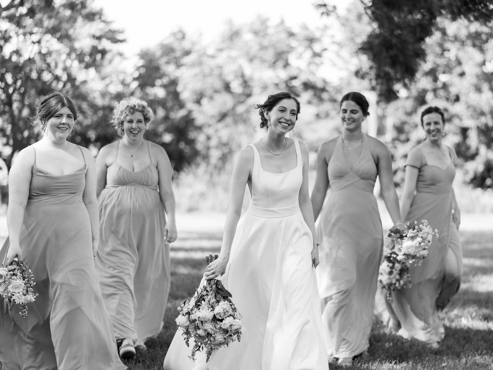 A wedding party in matching pale gray dresses walks through a garden holding bouquets in black and white photography.