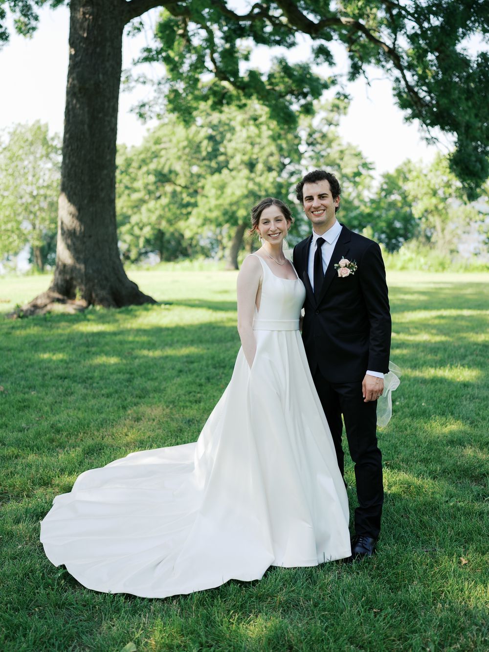 A bride in a flowing white wedding gown with a train and groom in black suit pose together on a sunny lawn under trees.