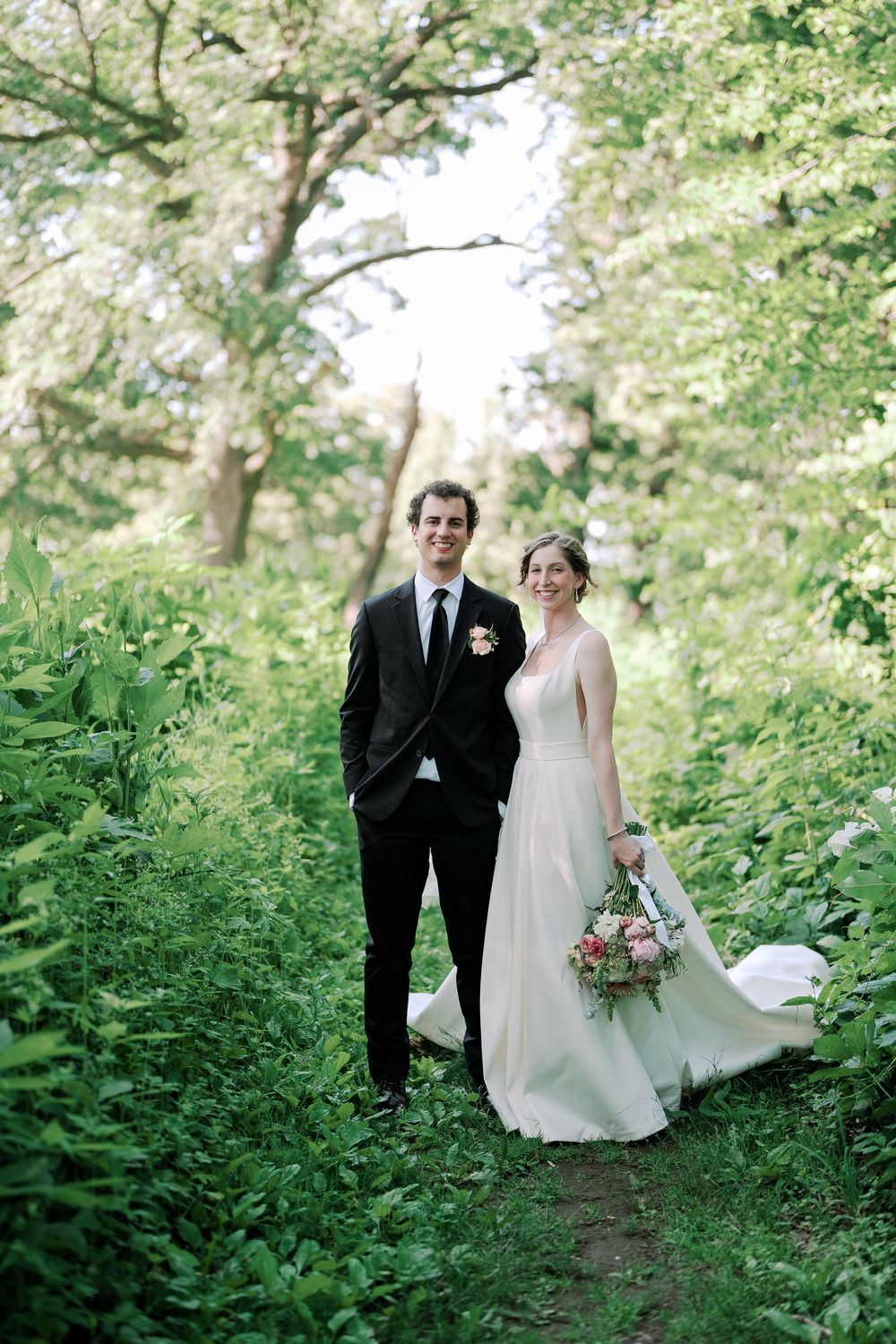 A couple in wedding attire stands on a verdant forest path surrounded by lush greenery and spring foliage.