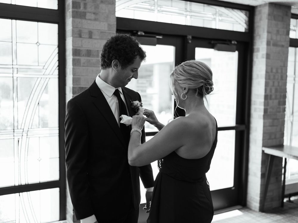 A tender moment at a formal event as someone adjusts the boutonniere on a suit jacket in black and white.