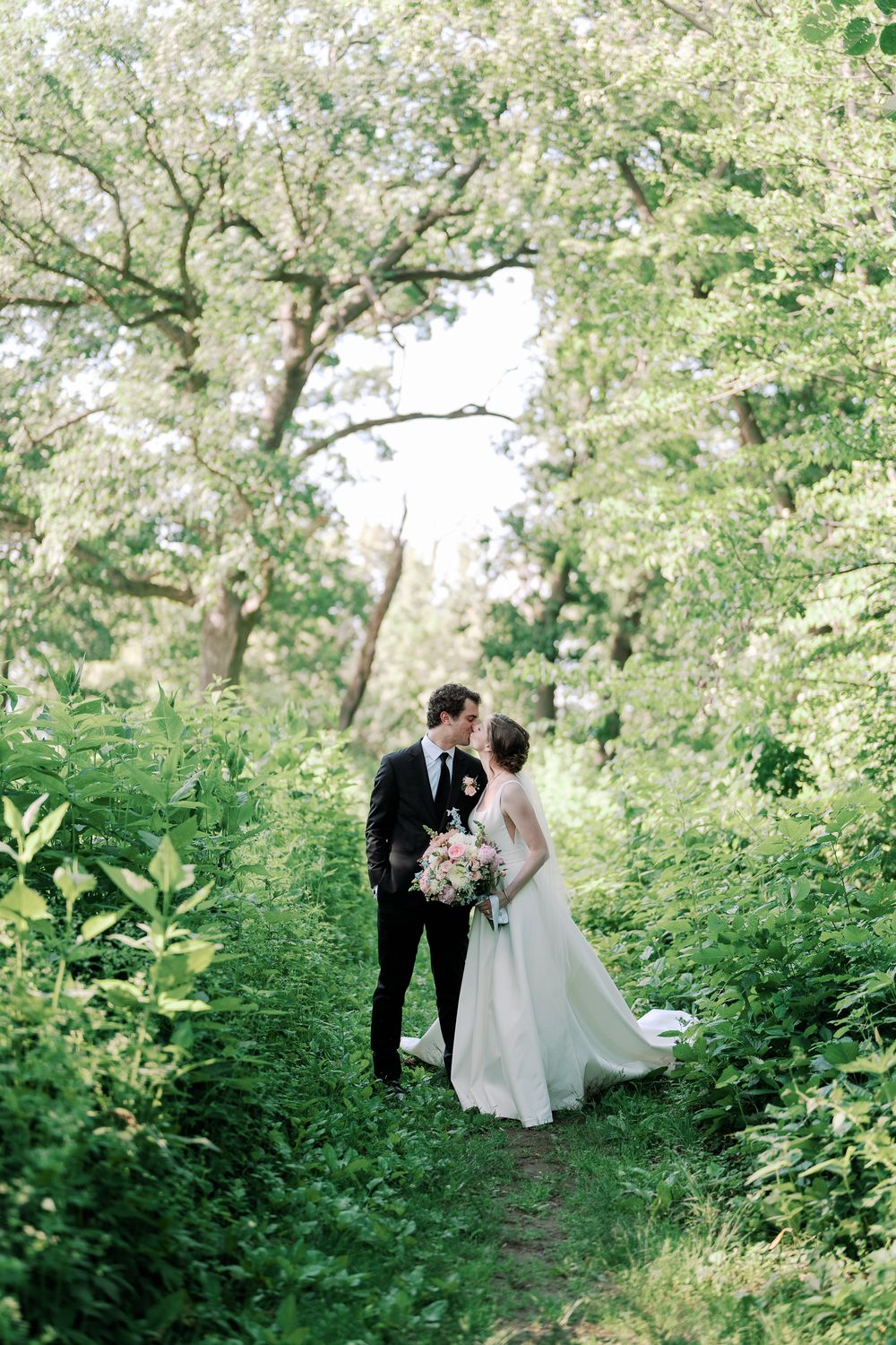 A romantic wedding couple standing on a garden path surrounded by lush greenery and blooming trees in spring.