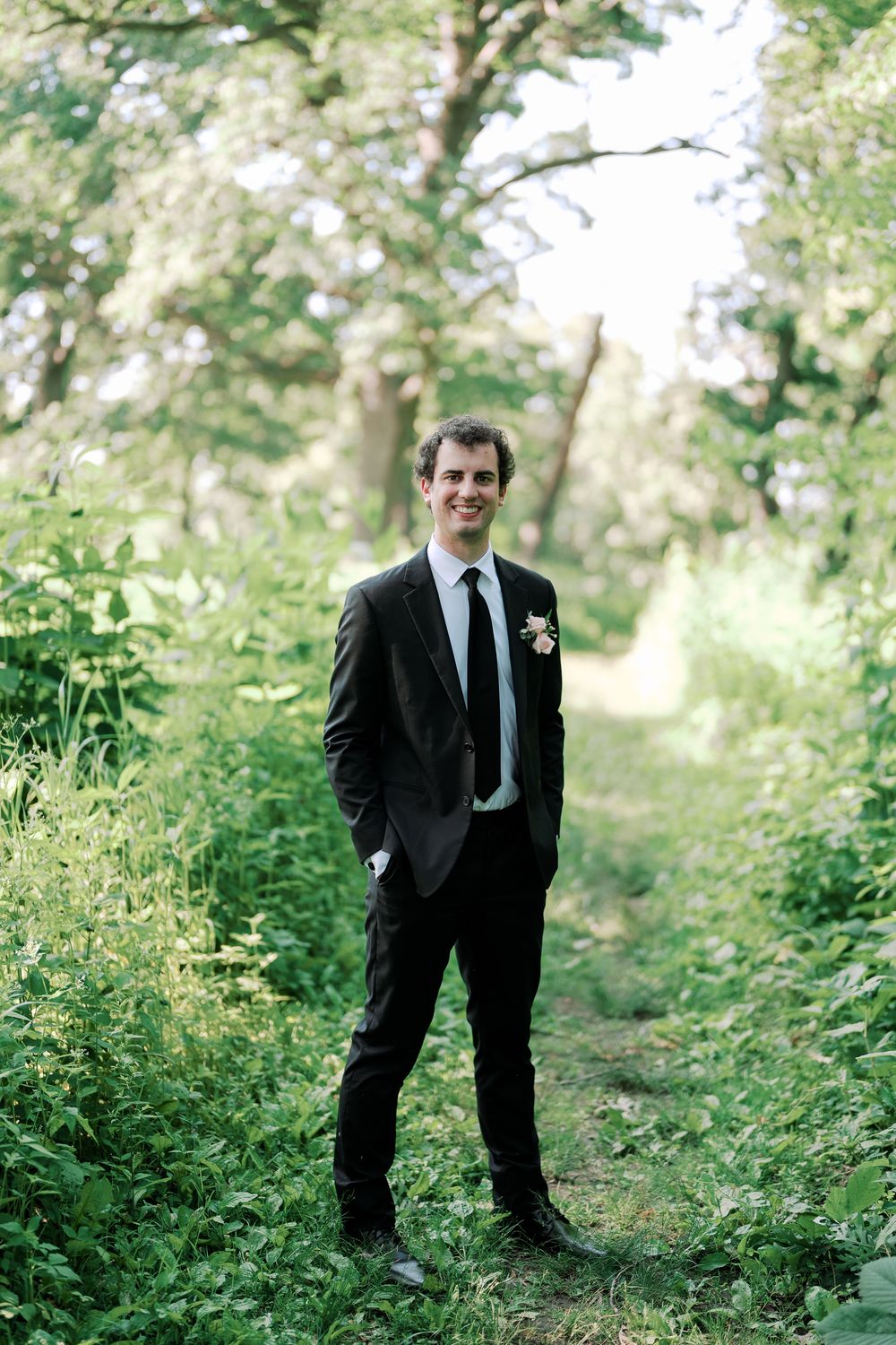 A groom in a dark suit stands outdoors on a wooded path surrounded by lush greenery and soft natural lighting.