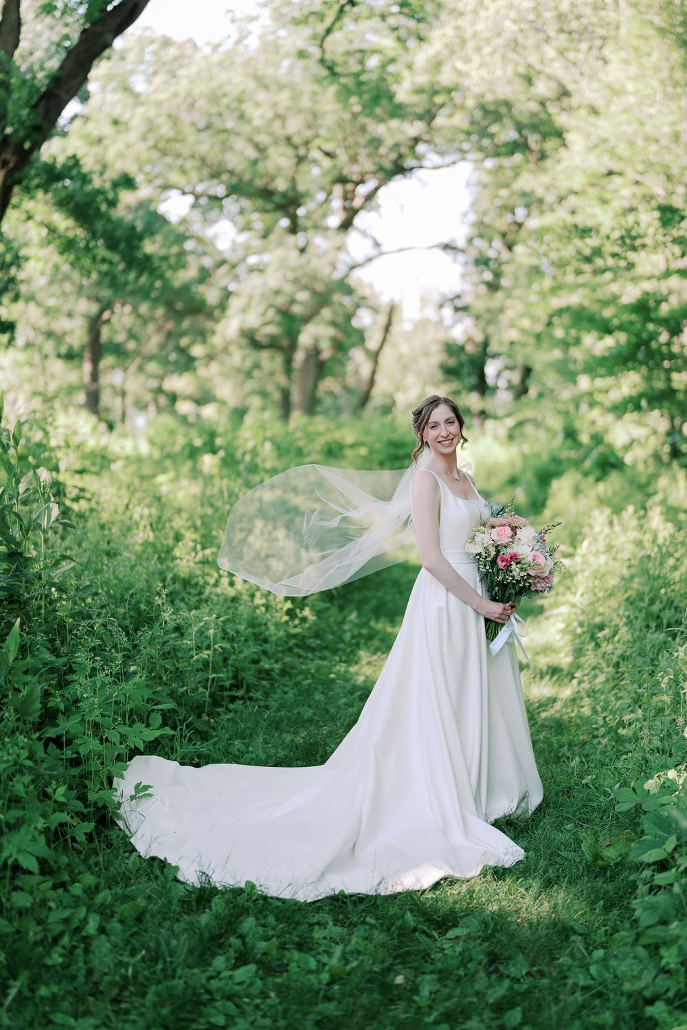 A bride in a flowing white wedding dress poses elegantly in a lush green garden setting with a floral bouquet.
