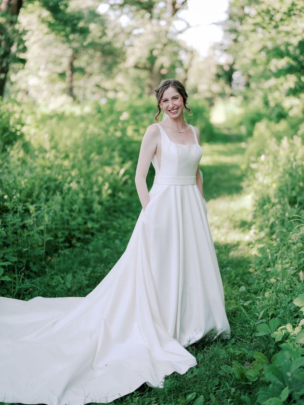 Elegant white wedding dress with pockets and long train photographed in a lush green outdoor setting.