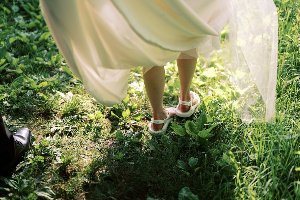 White dress hem and feet with strappy sandals standing on grass in soft sunlit garden setting.