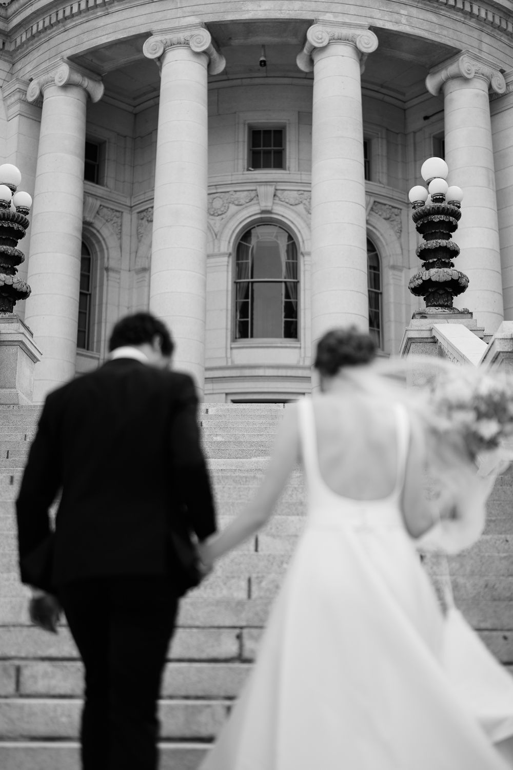 A bride and groom walk up grand marble steps toward a neoclassical building with white columns and ornate architecture.