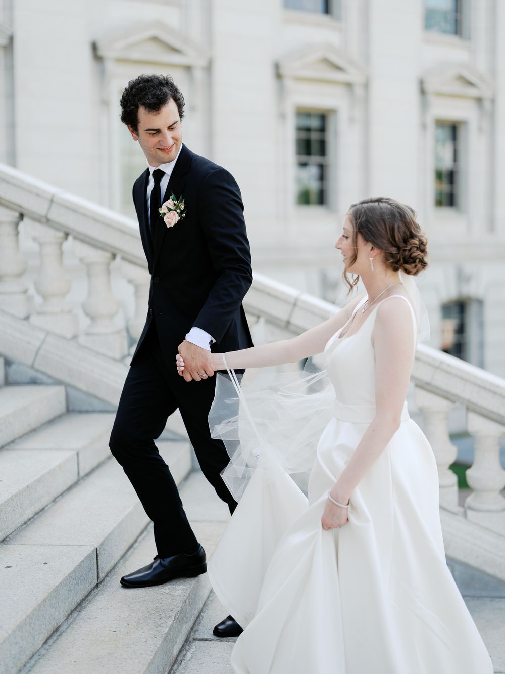 A couple in wedding attire demonstrates different poses while ascending elegant white marble stairs outdoors.