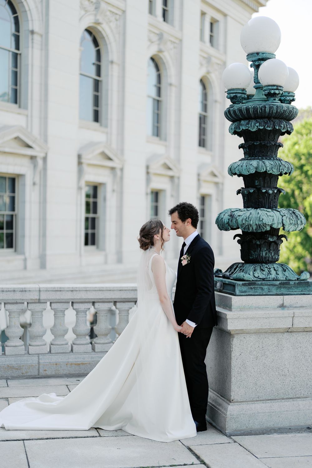 A bride and groom share an intimate moment by an ornate lamp post near a white stone building with arched windows.