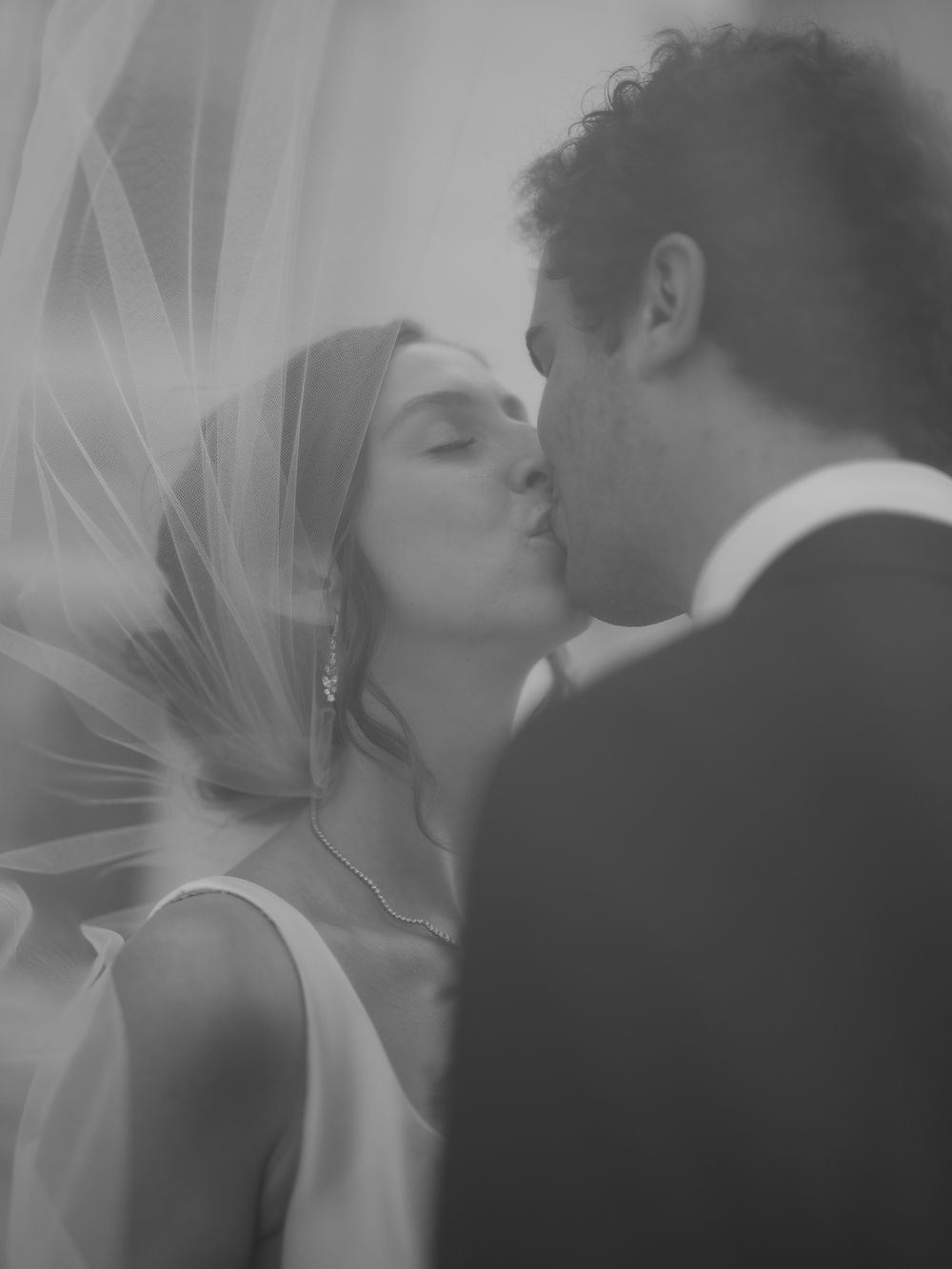 A romantic black and white wedding photo showing a couple sharing a tender kiss with a flowing veil in soft light.
