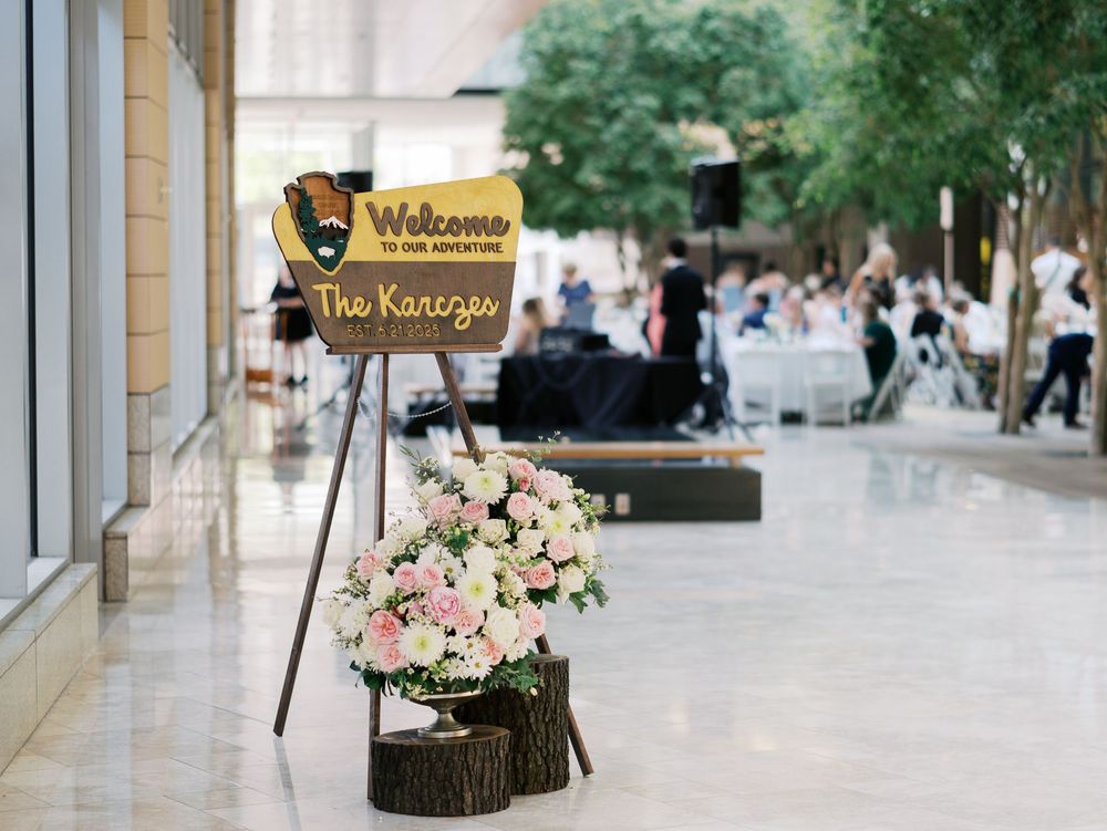 Welcome sign and floral arrangement on wood slices decorate a venue entrance with guests mingling in background.