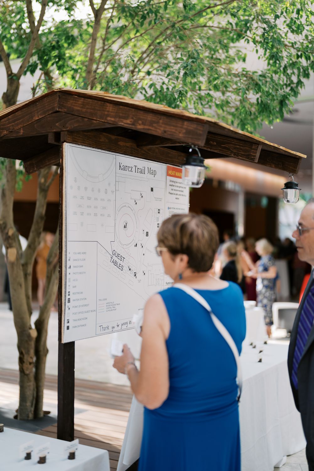 People gather around a posted board display with information under a wooden overhang at an outdoor venue.