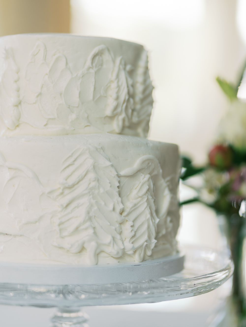 Close-up view of a white textured wedding cake with rustic frosting details on a glass cake stand.