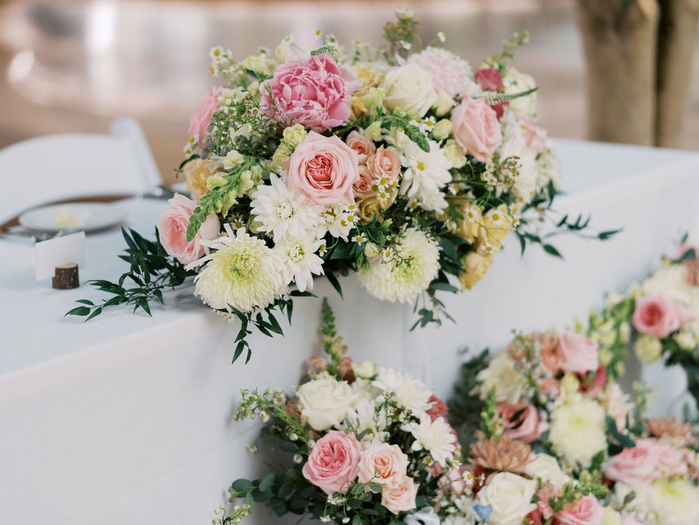 Elegant floral centerpiece with blush pink roses, white dahlias and greenery on white tablecloth for wedding reception.