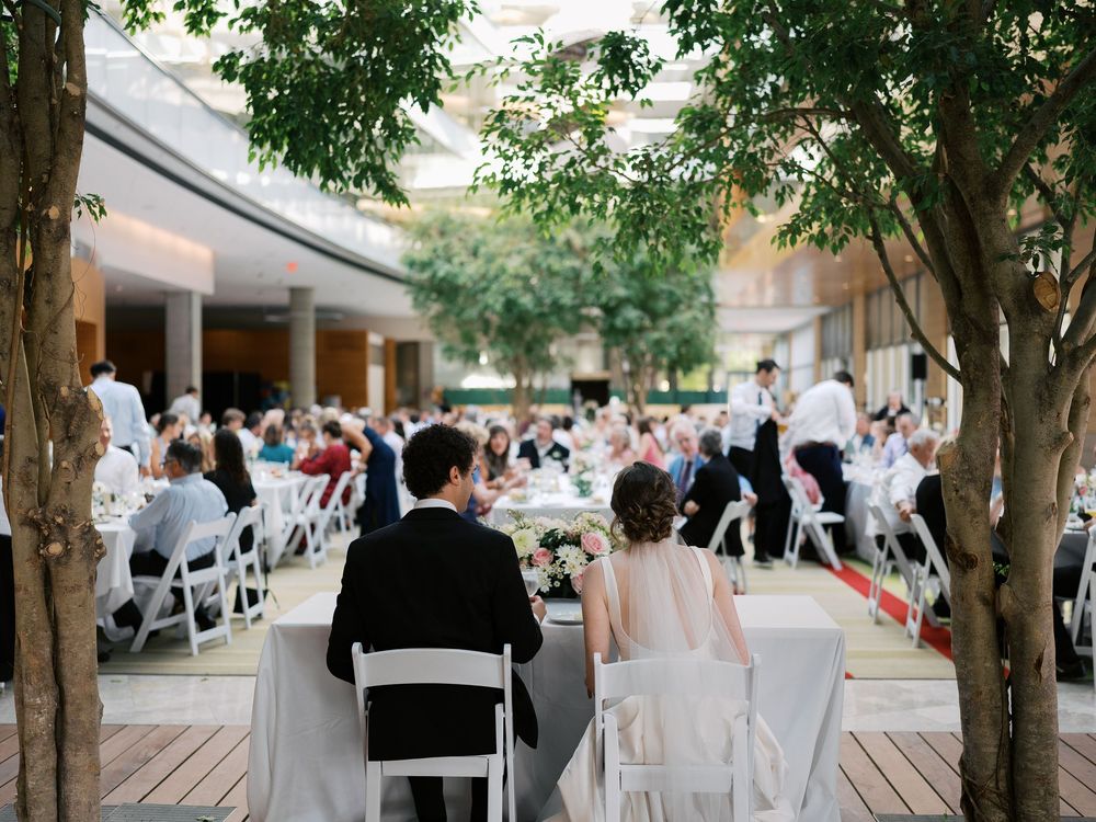 Outdoor wedding ceremony setup under trees with white chairs and guests seated in a courtyard space.