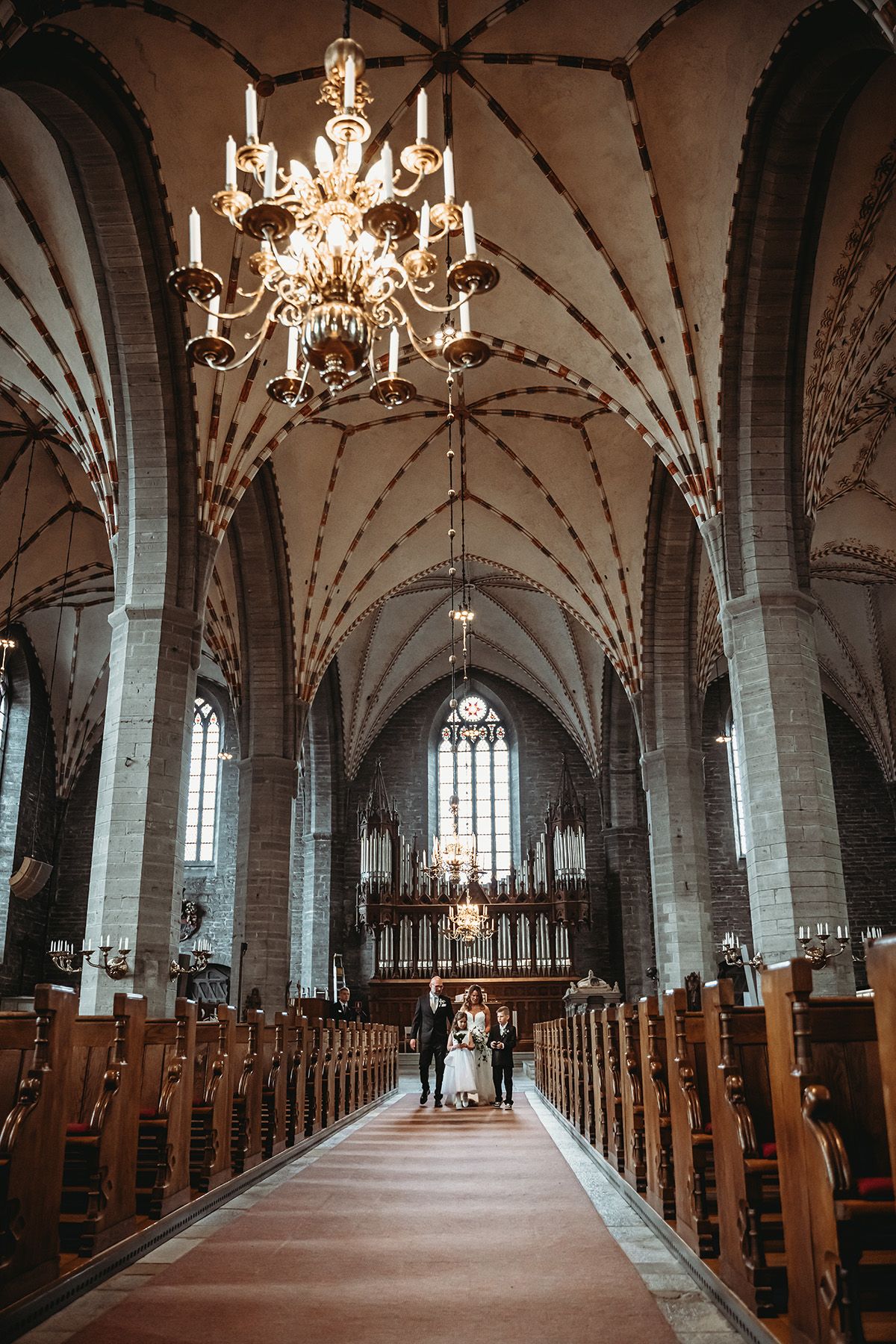 Vigsel i Klosterkyrkan i Vadstena, brudparet går med sina brudnäbbar uppför altargången och man ser det välvda taket ovanför dem.
