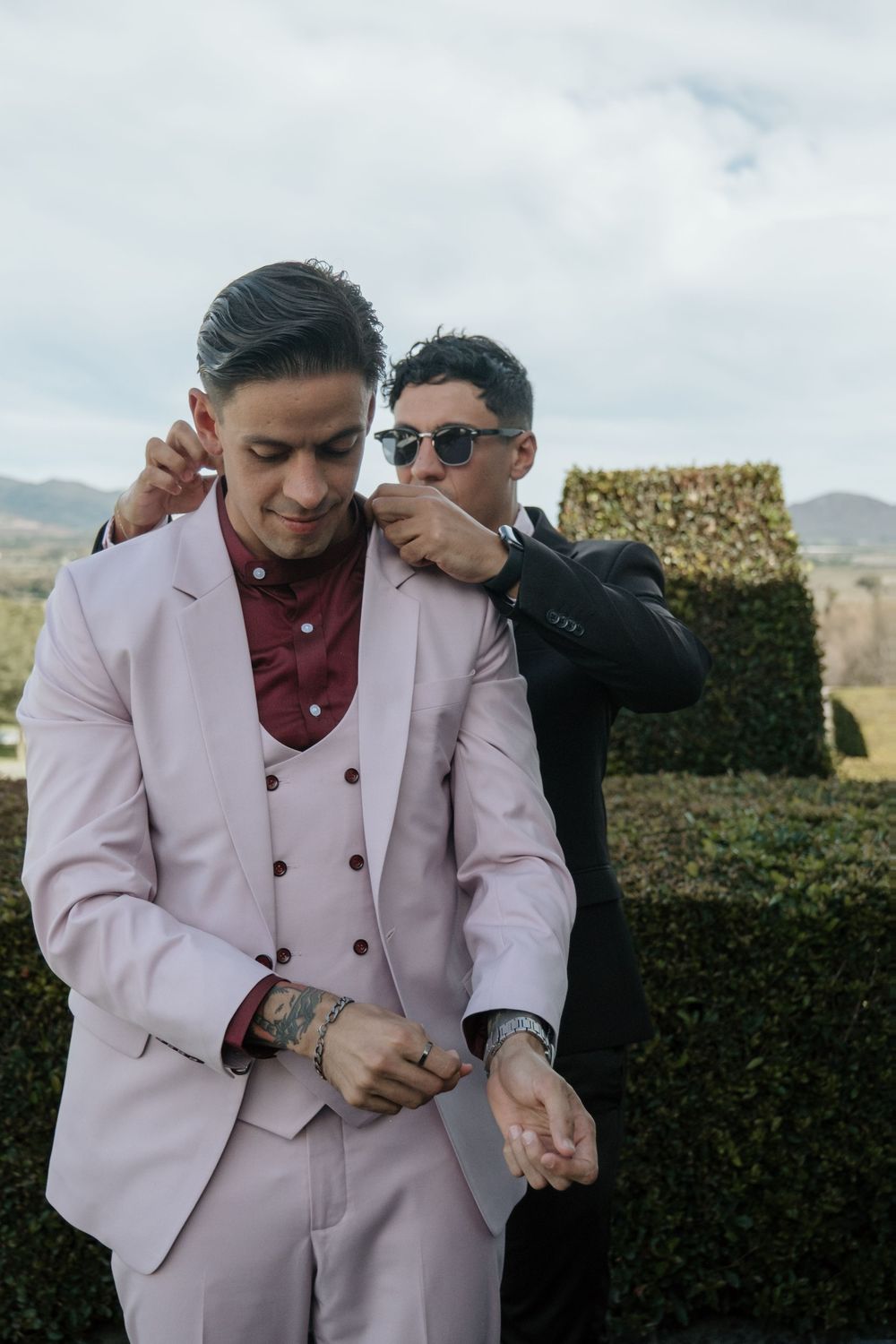 A groom dressed in a pink double-breasted suit shares a lighthearted moment with the best man in an outdoor garden setting.