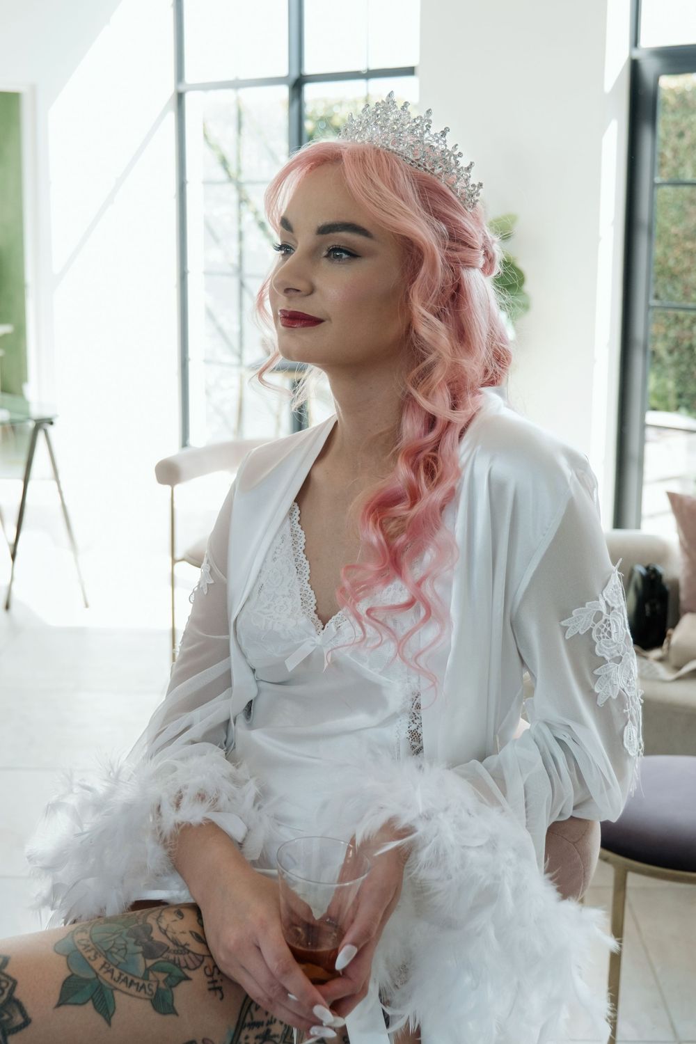 A person with pink wavy hair wearing a white robe and tiara sits in a bright room near windows.