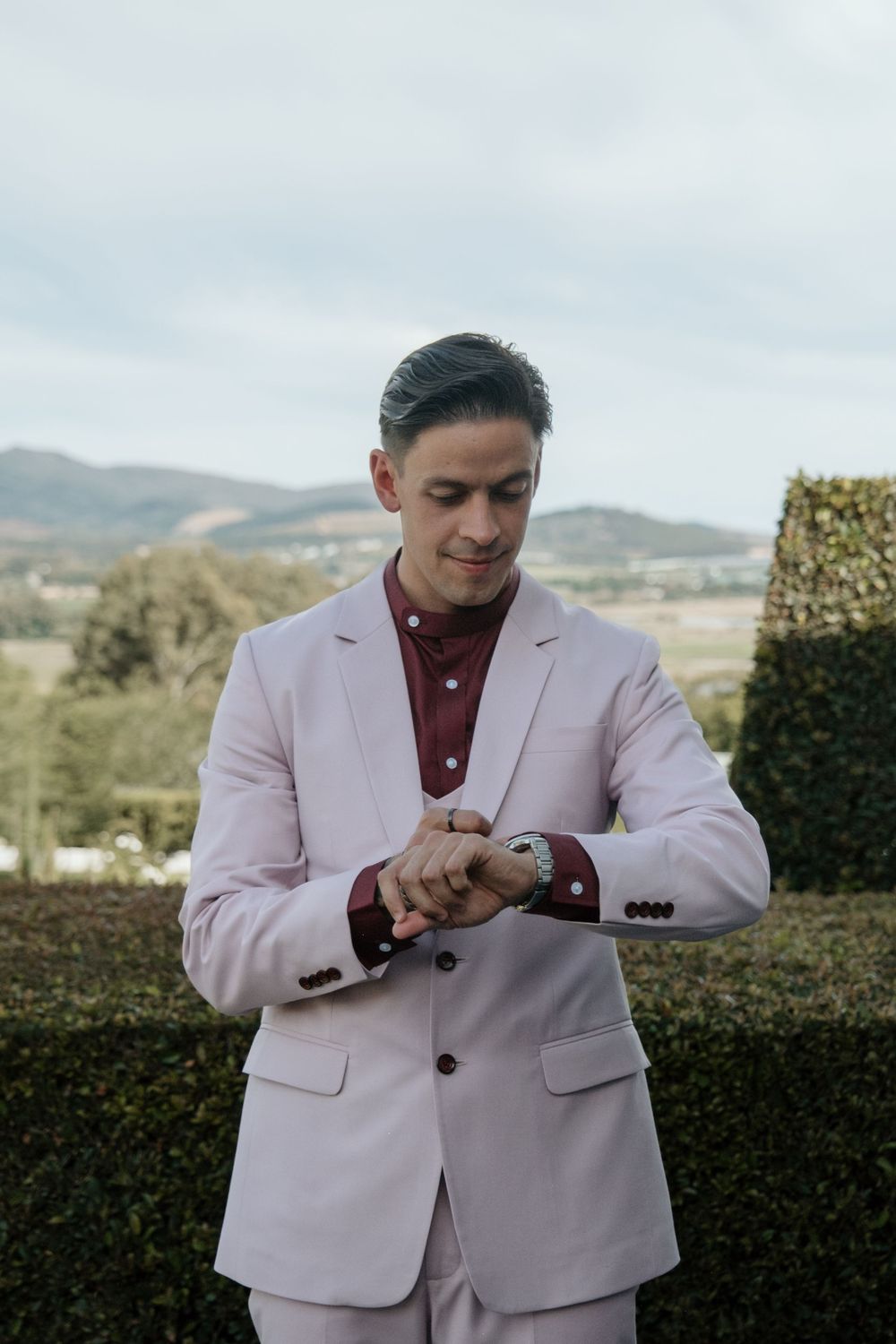 Someone in a light pink suit and burgundy shirt checks their watch against an outdoor mountain landscape.