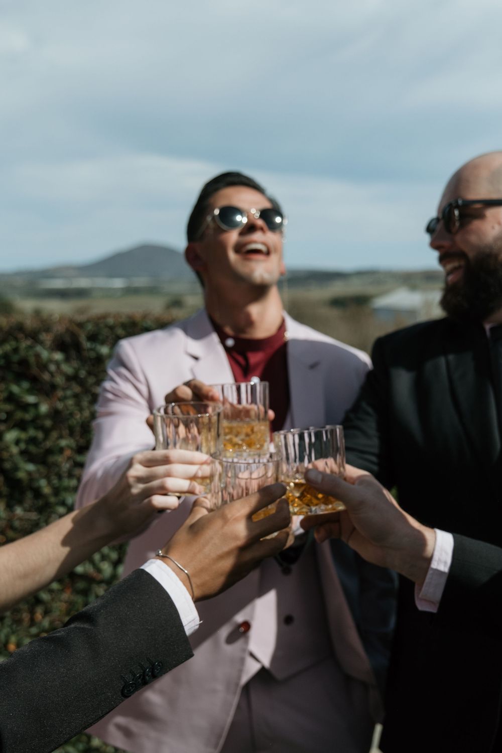 Friends in formal attire toast with whiskey glasses outdoors against a scenic mountain backdrop.