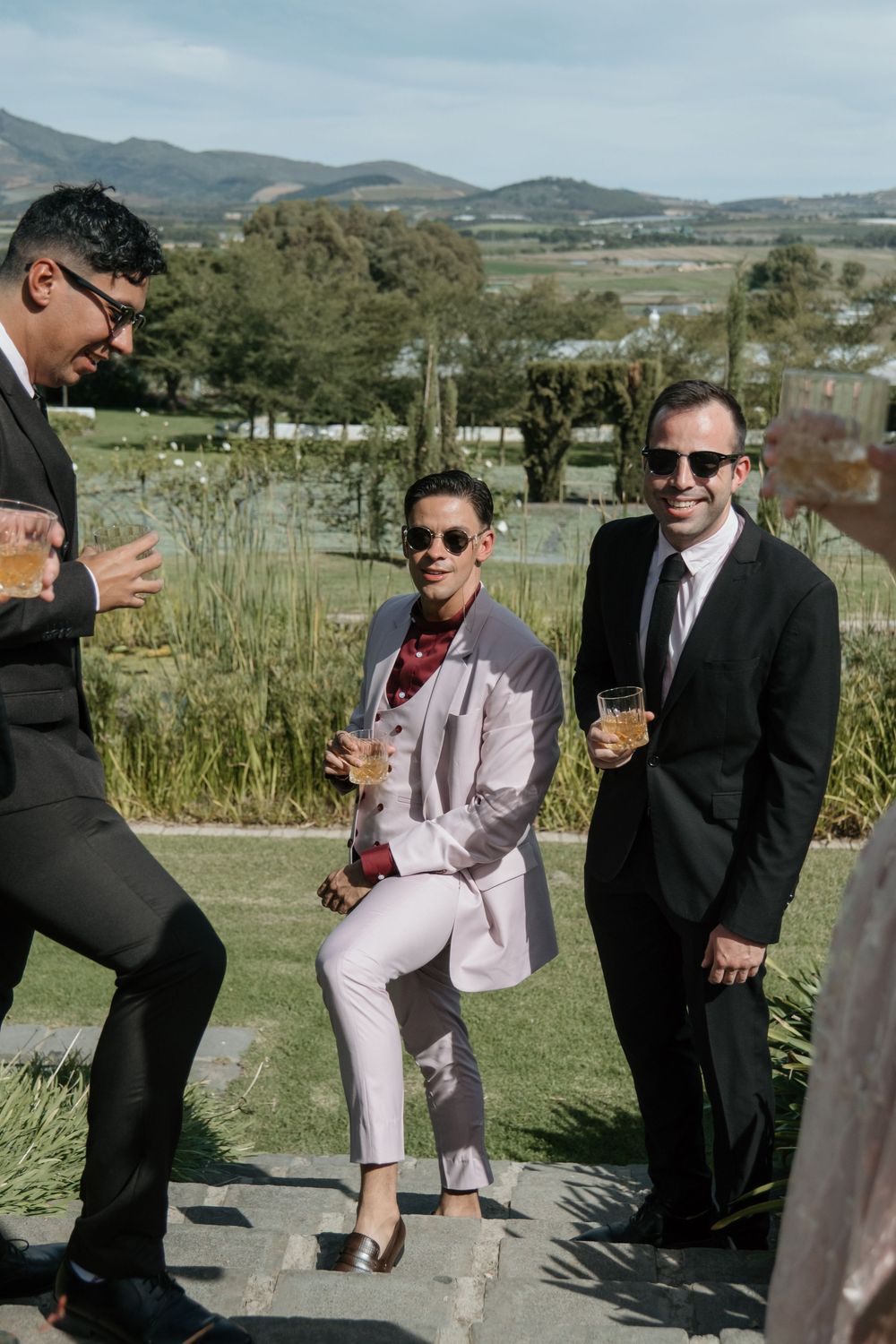 Guests in formal attire enjoying drinks and conversation on a scenic hillside during an outdoor event.