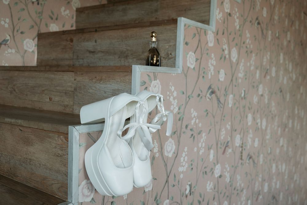 White ballet shoes hang on wooden stairs next to a decorative bottle against floral wallpaper.