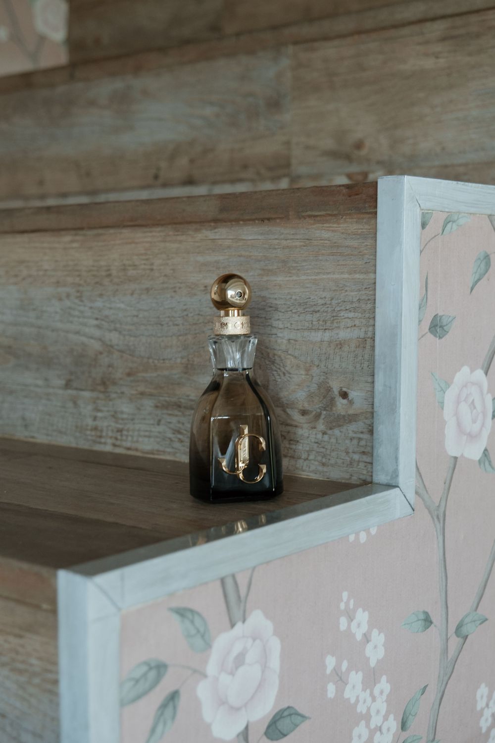 Vintage smoky glass decanter with gold stopper sits on wooden shelf beside floral patterned white frame.