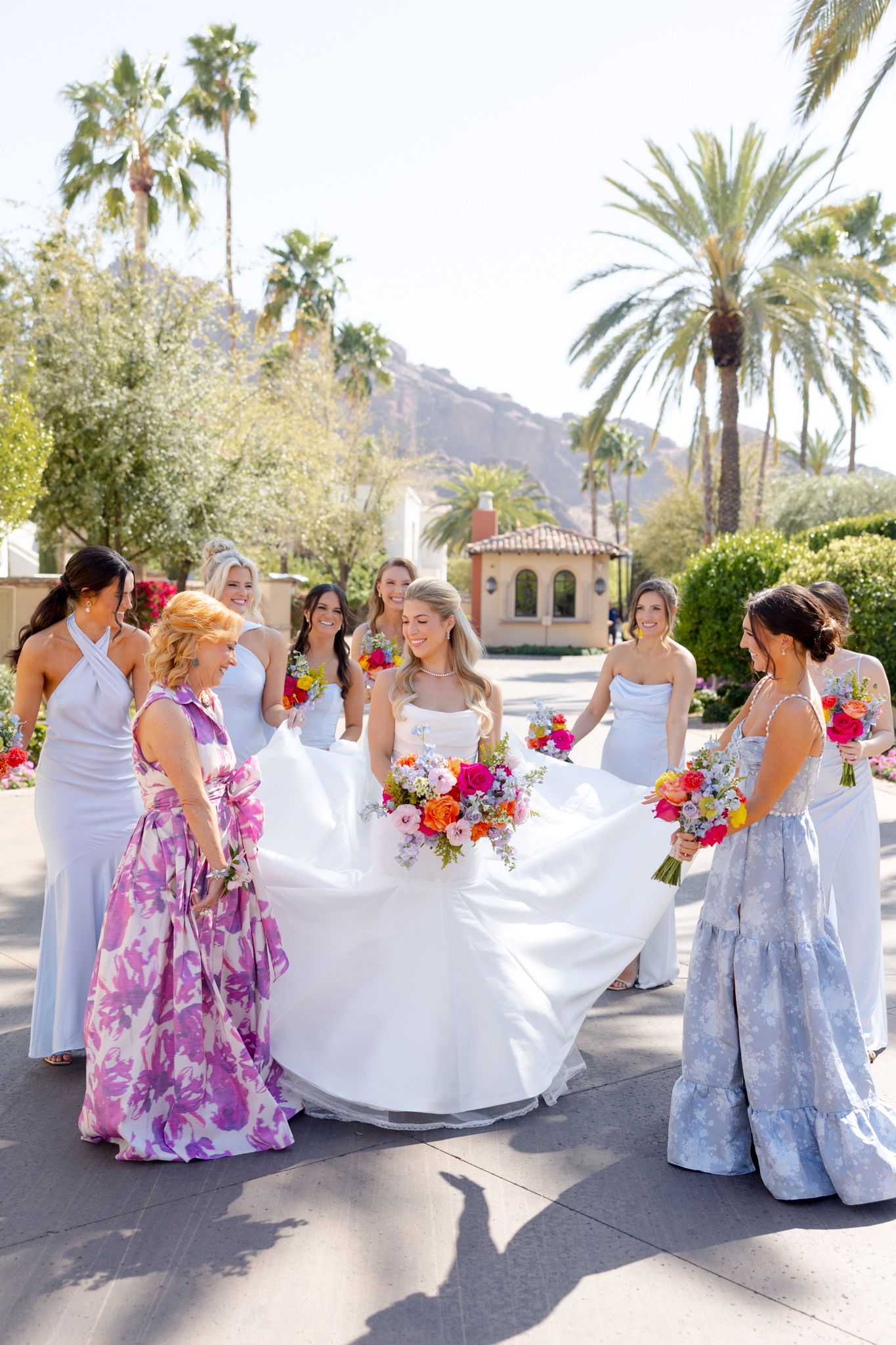 Bridal party in colorful floral dresses and white gowns gather on palm-lined path with vibrant bouquets at outdoor venue.
