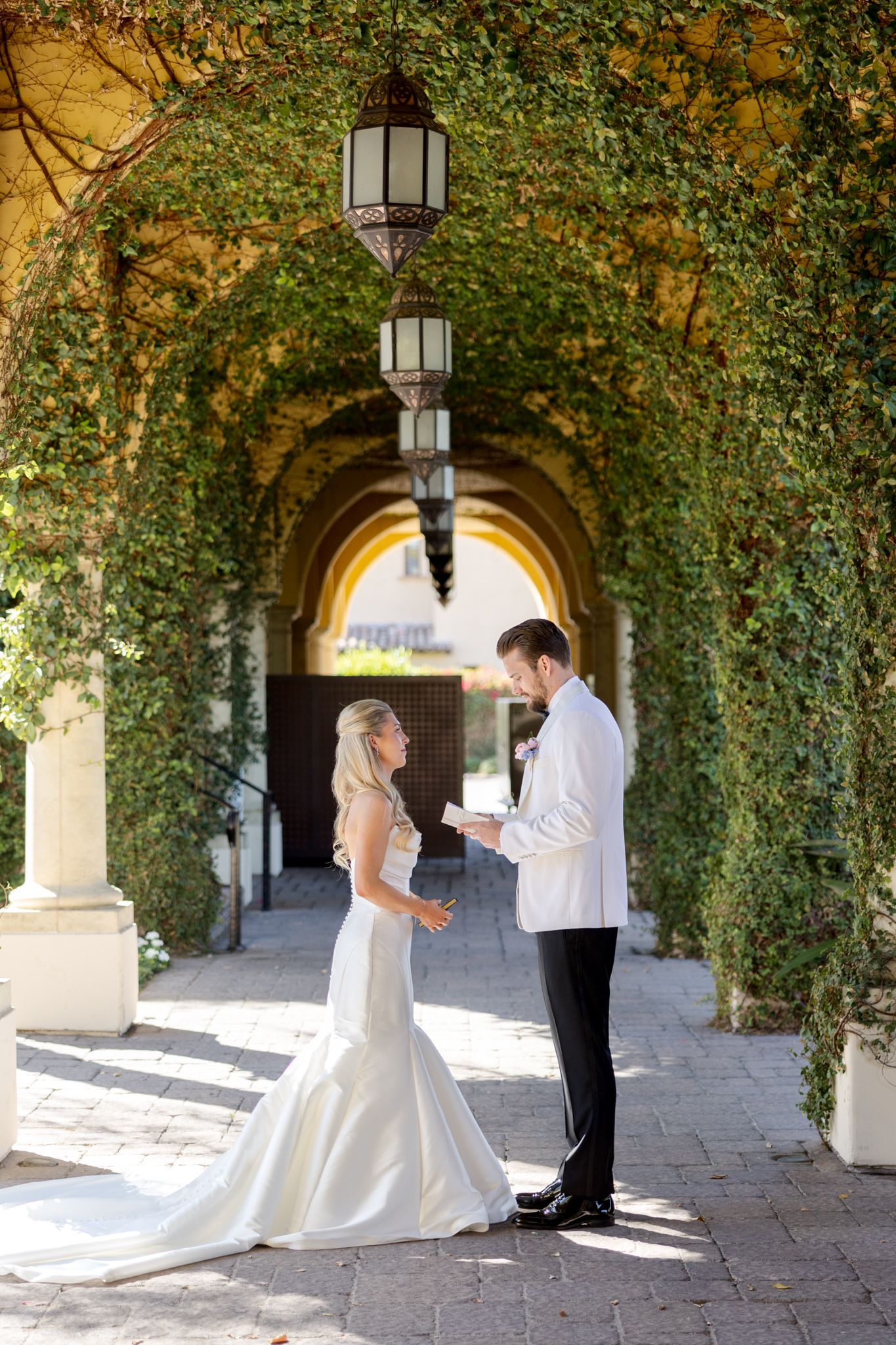 Couple exchanges vows beneath ivy-covered stone archway with hanging lanterns and golden sunlight streaming through.