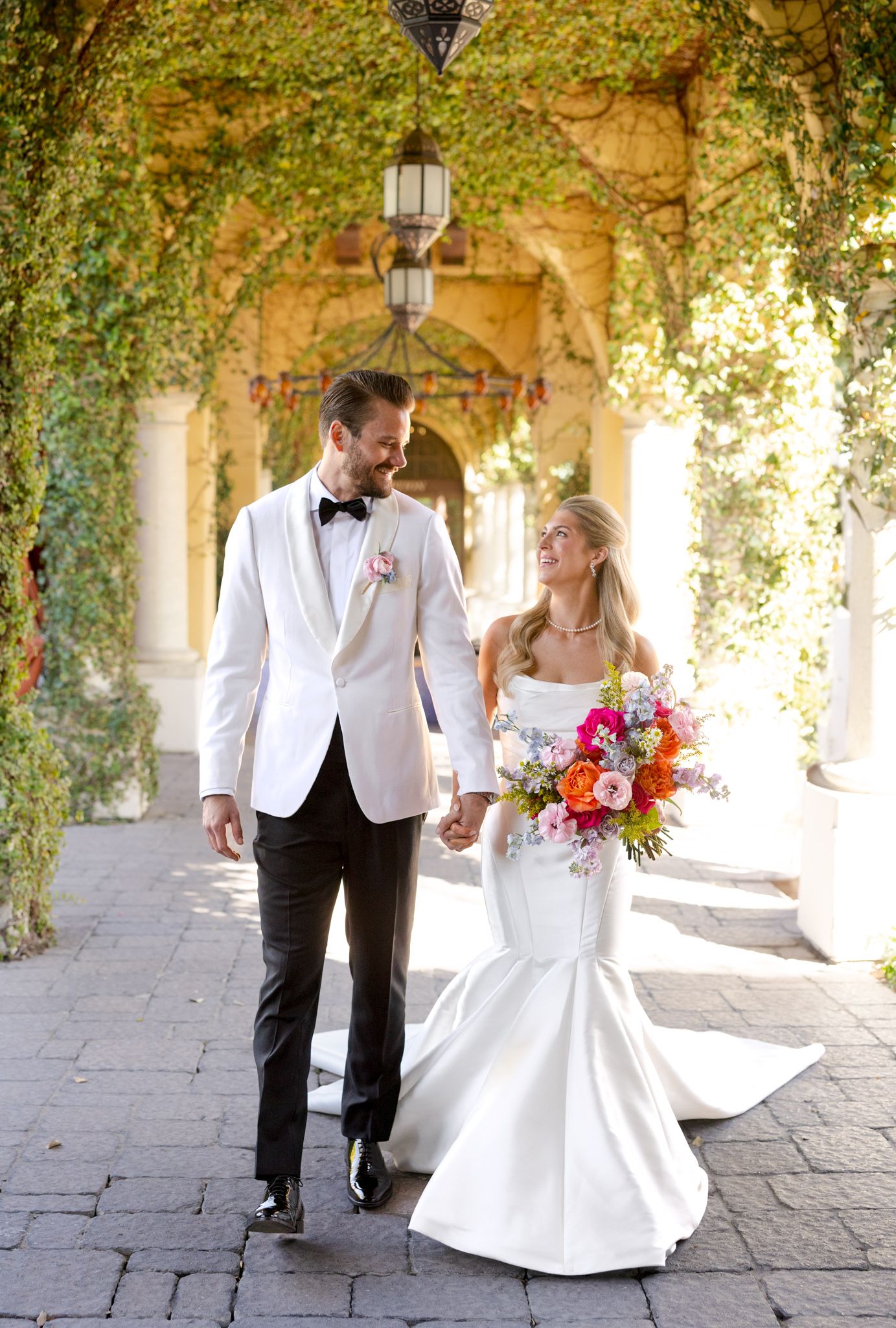 Newlyweds walking through elegant covered walkway with hanging vines and lanterns, bride holding colorful bouquet.