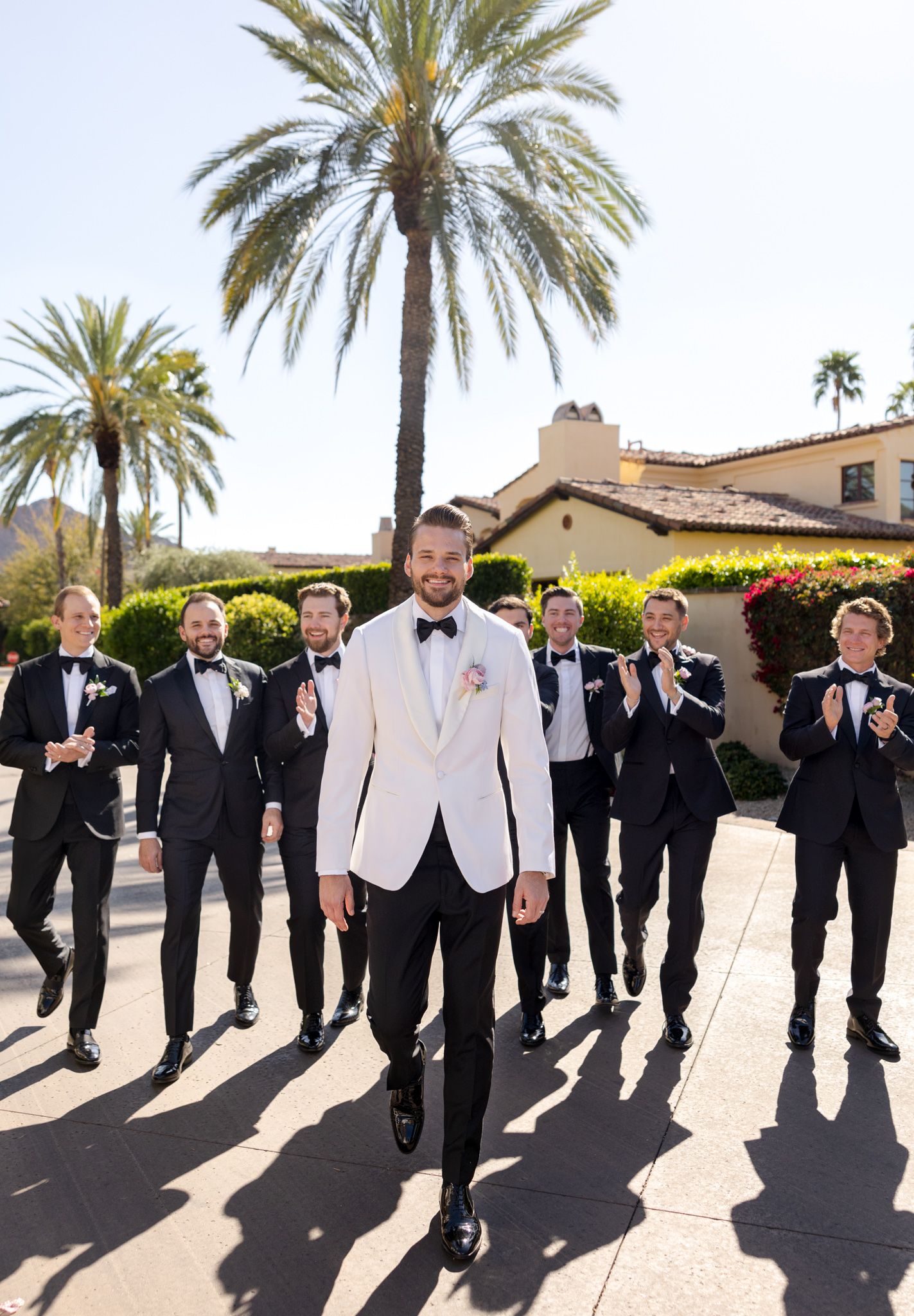 Wedding party in formal attire walking together at a luxurious venue with palm trees and Spanish-style architecture.