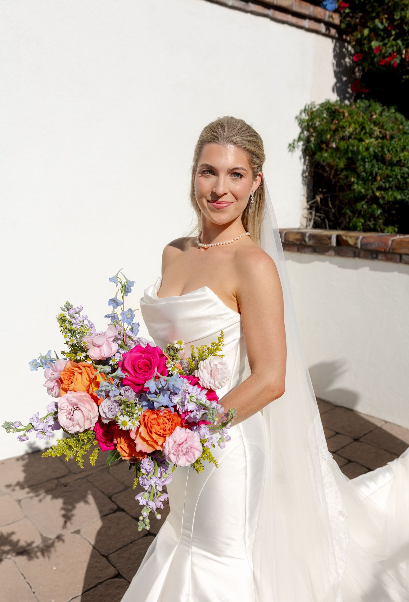 A bride in a strapless white gown holds a vibrant bouquet of pink, orange and purple flowers against a white wall.