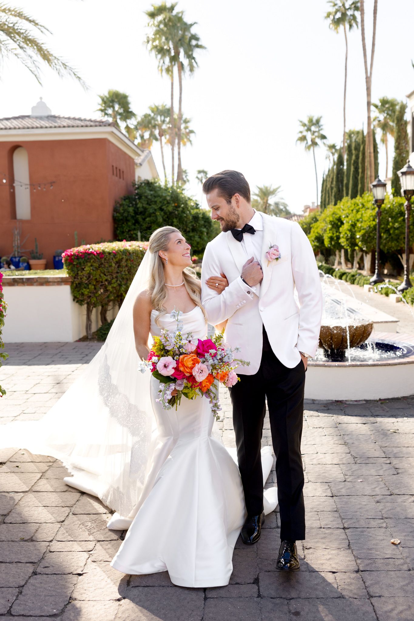 A wedding couple walks together in a palm-tree lined courtyard, the bride holding a vibrant colorful bouquet.