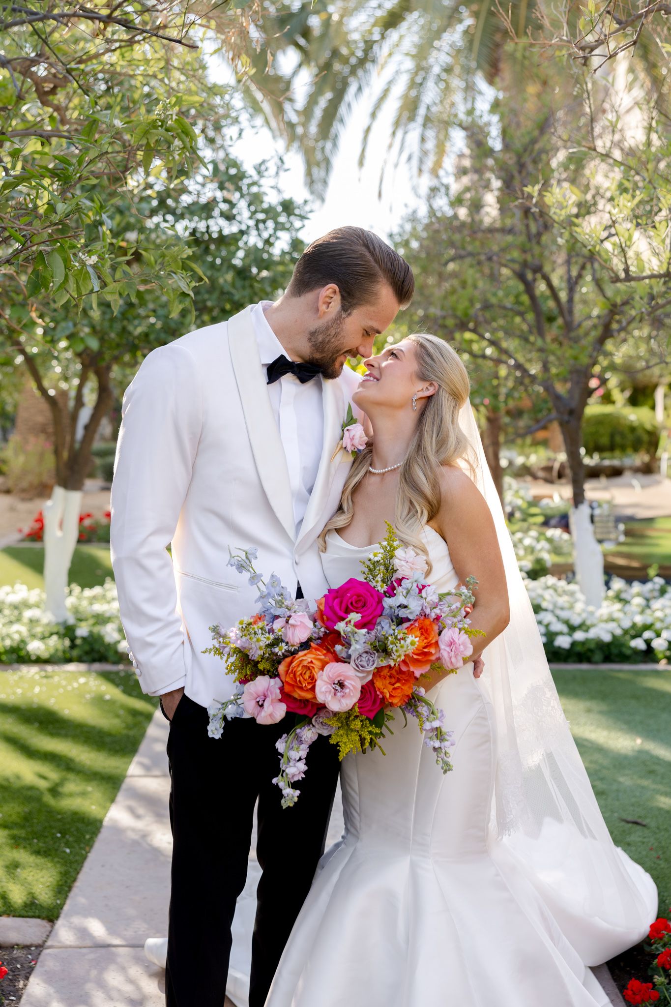 A couple shares a romantic moment in a garden setting with palm trees and colorful floral bouquet.