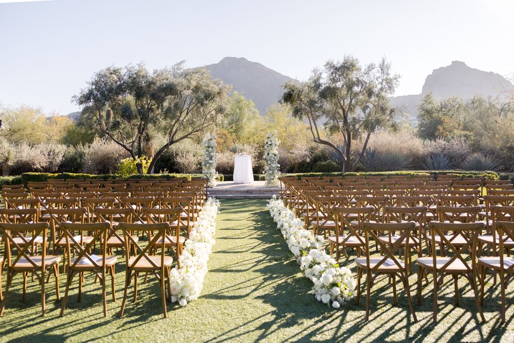 Rustic wooden chairs line a grassy aisle leading to a white arch for an outdoor wedding ceremony with mountain views.