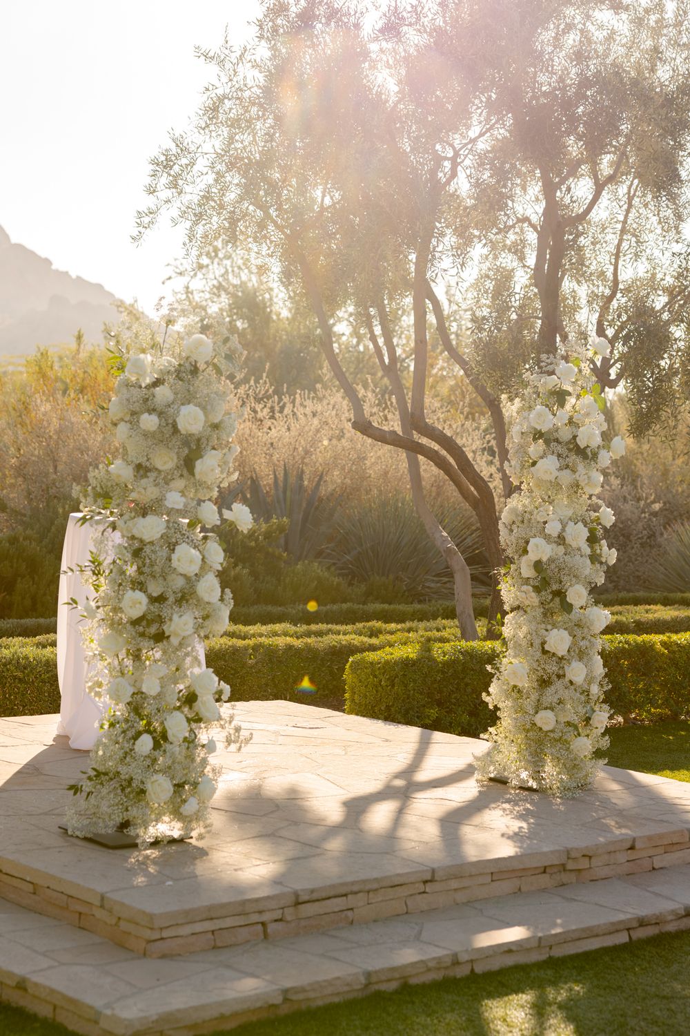 Elegant white floral archway setup in a garden during golden hour sunset creates a romantic outdoor wedding ceremony backdrop.